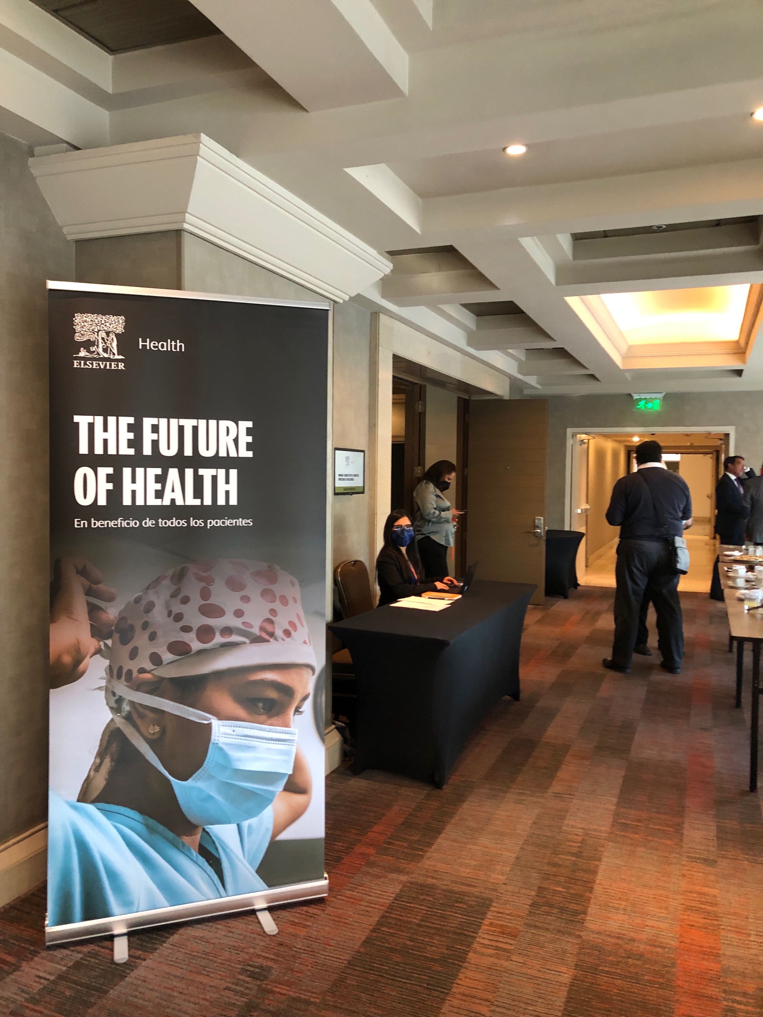 Fotografía de la recepción del evento organizado por Elsevier. Se ve un pendón con la información y algunos asistentes de espalda. 