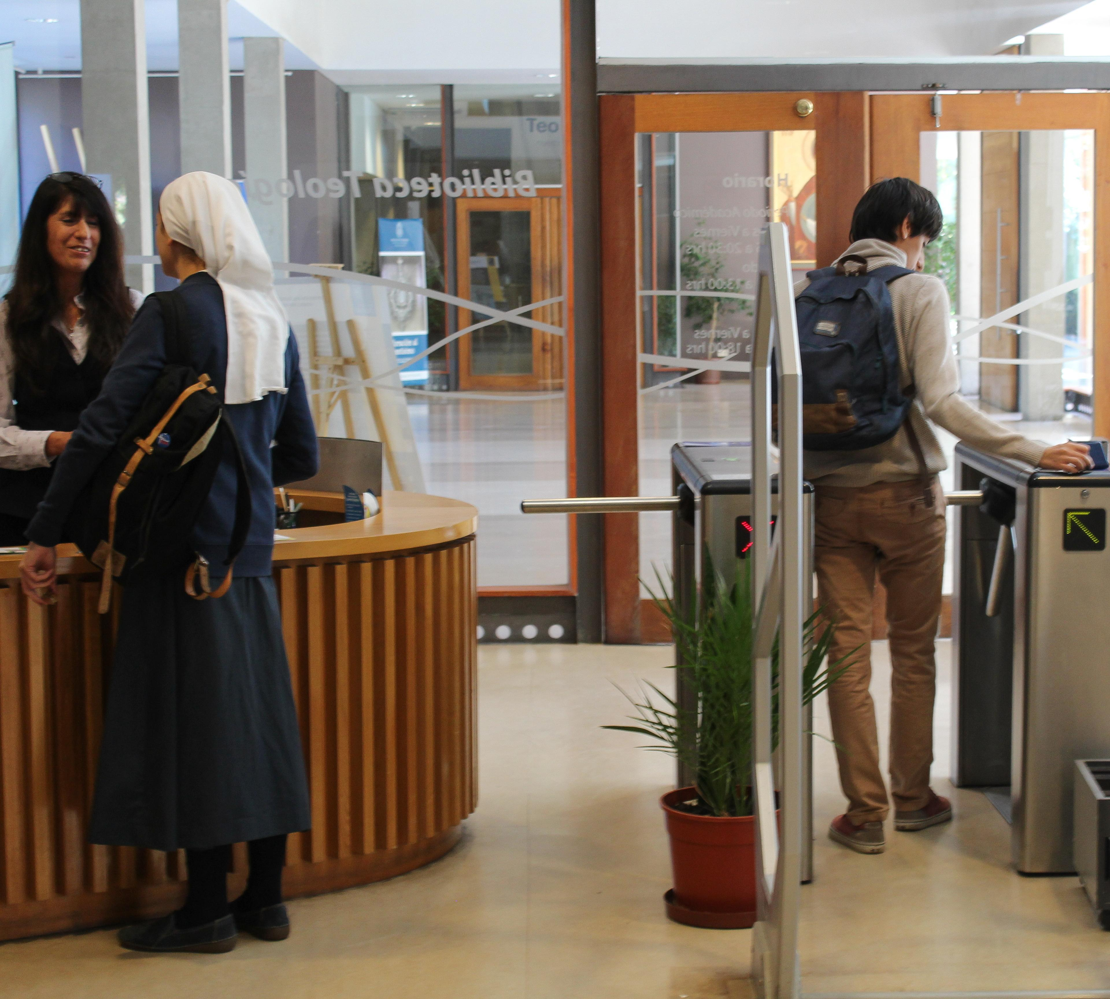 Fotografía de la entrada de la Biblioteca de Teología. Se ve una religiosa consultando en el mesón y un estudiante pasando por el acceso.