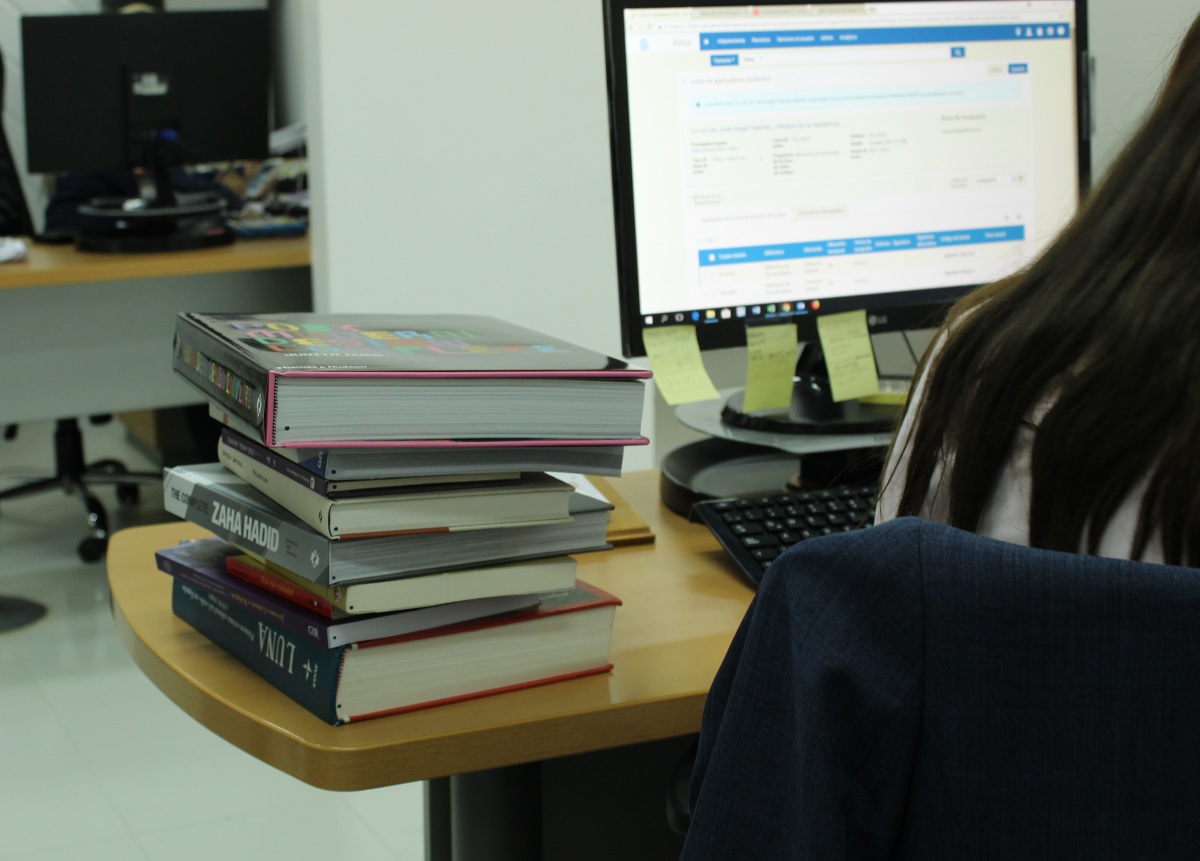 Fotografía de una oficina interna de Bbliotecas UC. En primer plano se ve una torre de libros  junto a un puesto de una persona con un computador. Esta fotografía muestra cómo se lleva el cabo el proceso de adquisición, registro y digitalización de los libros.