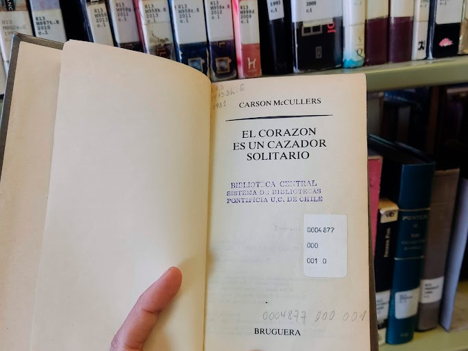 Fotografía del libro "El corazón es un cazador solitario" abierto en la primera página. La foto está sacada frente a una estantería de libros de Bibliotecas UC.